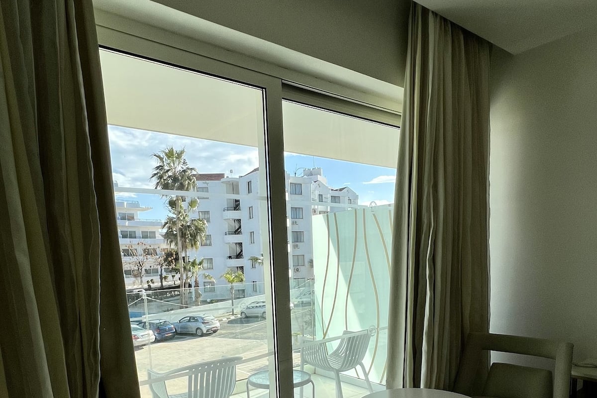 Ett modernt hotellrum med en stor skjutdörr i glas som leder ut till en balkong med utsikt över en strand och byggnader. På balkongen finns ett runt bord och två stolar.