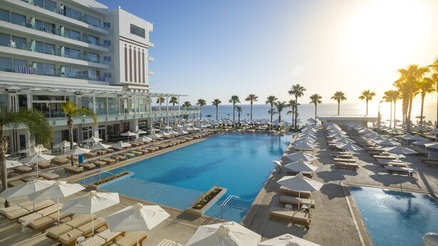 A hotel pool area with multiple sun loungers and umbrellas, a clear view of the ocean, and a modern building with balconies. - Constantinos The Great Beach Hotel (Paralimni)