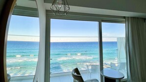 A modern hotel room with a large window offering a view of the ocean. There is a round mirror, a chair, and a small table. - Constantinos The Great Beach Hotel (Paralimni)