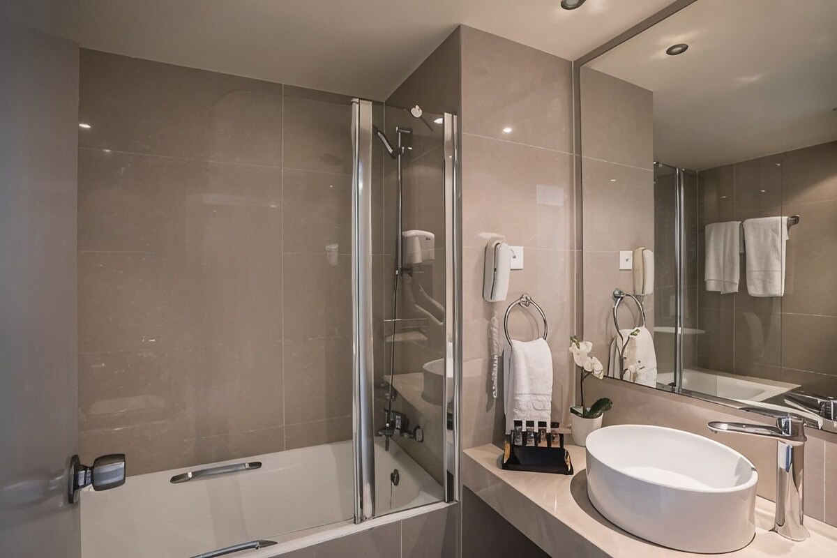 A modern bathroom with a glass-enclosed shower, a large sink, and a mirror reflecting a towel rack and a potted plant.