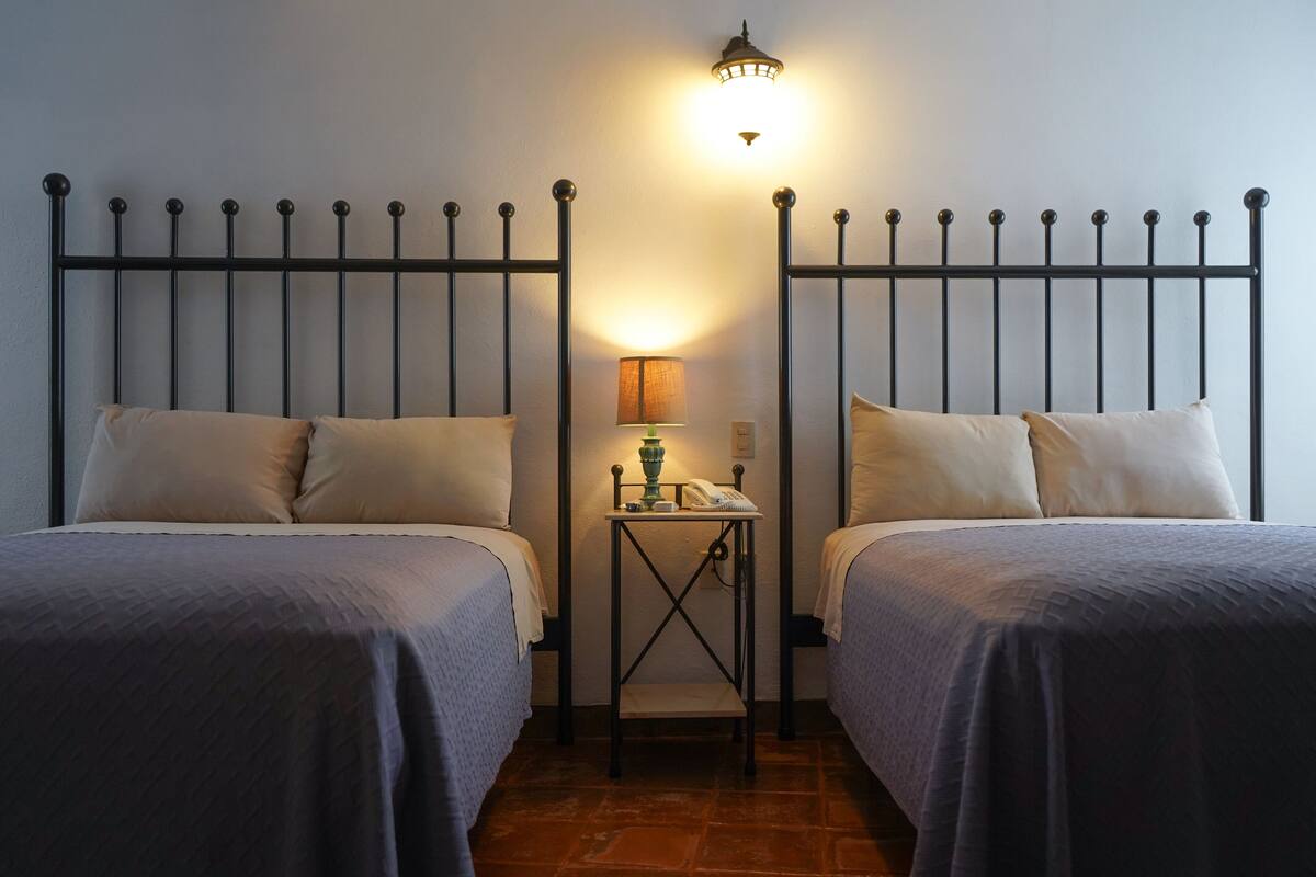 Una habitació amb dos llits, una tauleta de nit amb una làmpada i una làmpada de paret.