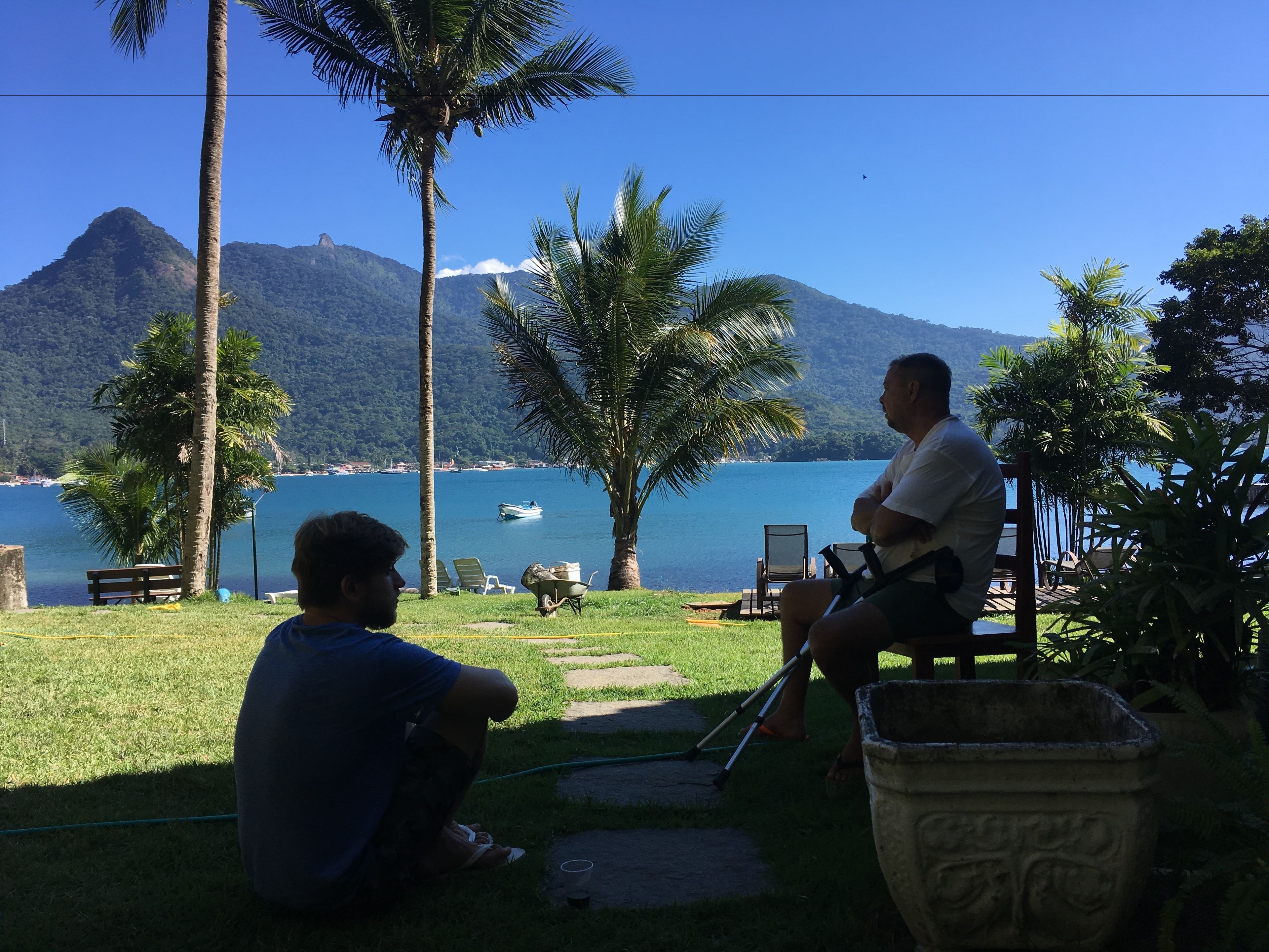 Pátio com duas pessoas sentadas, vista para um lago com barco e montanhas ao fundo.
