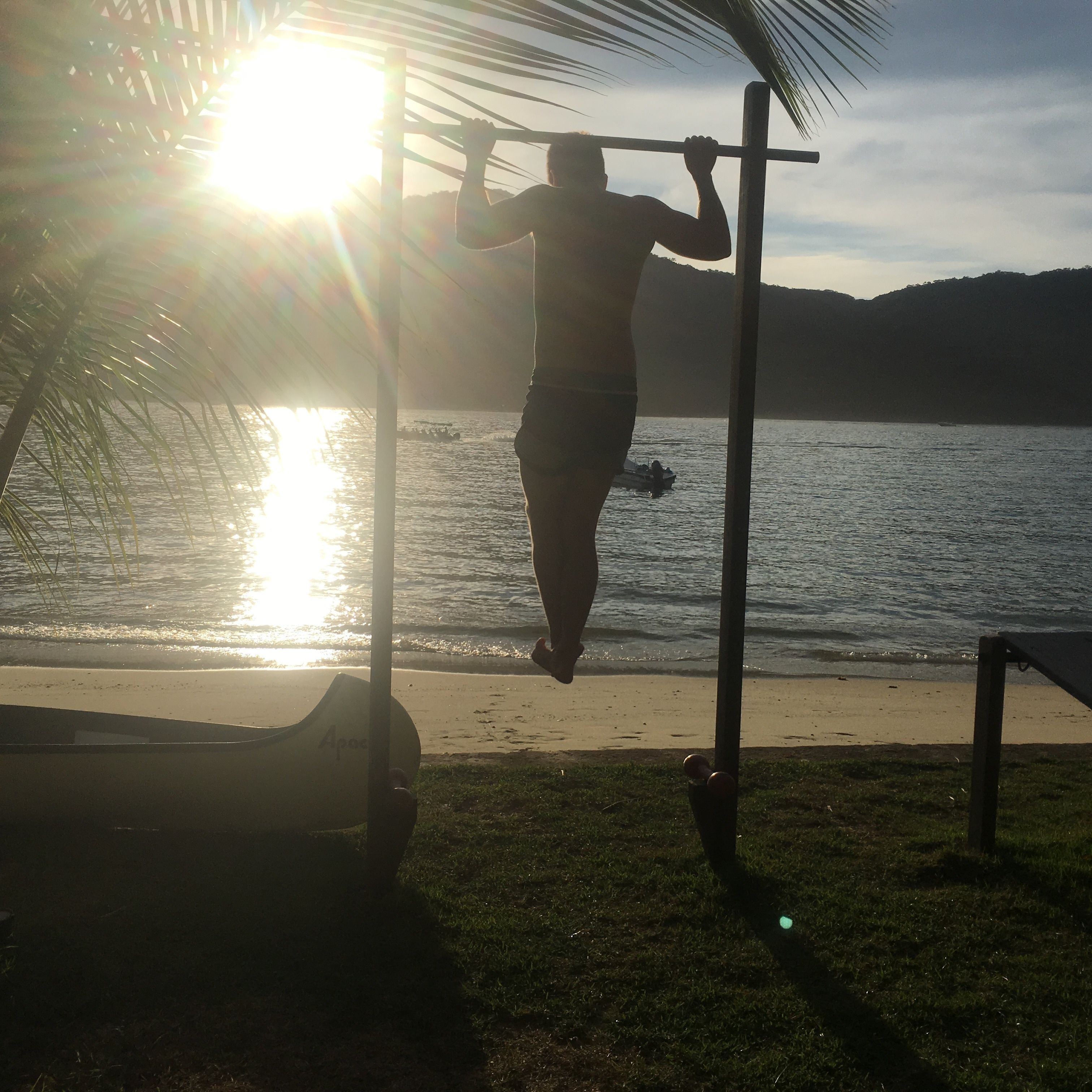 A person doing pull-ups on a bar by a lake with a boat in the background.