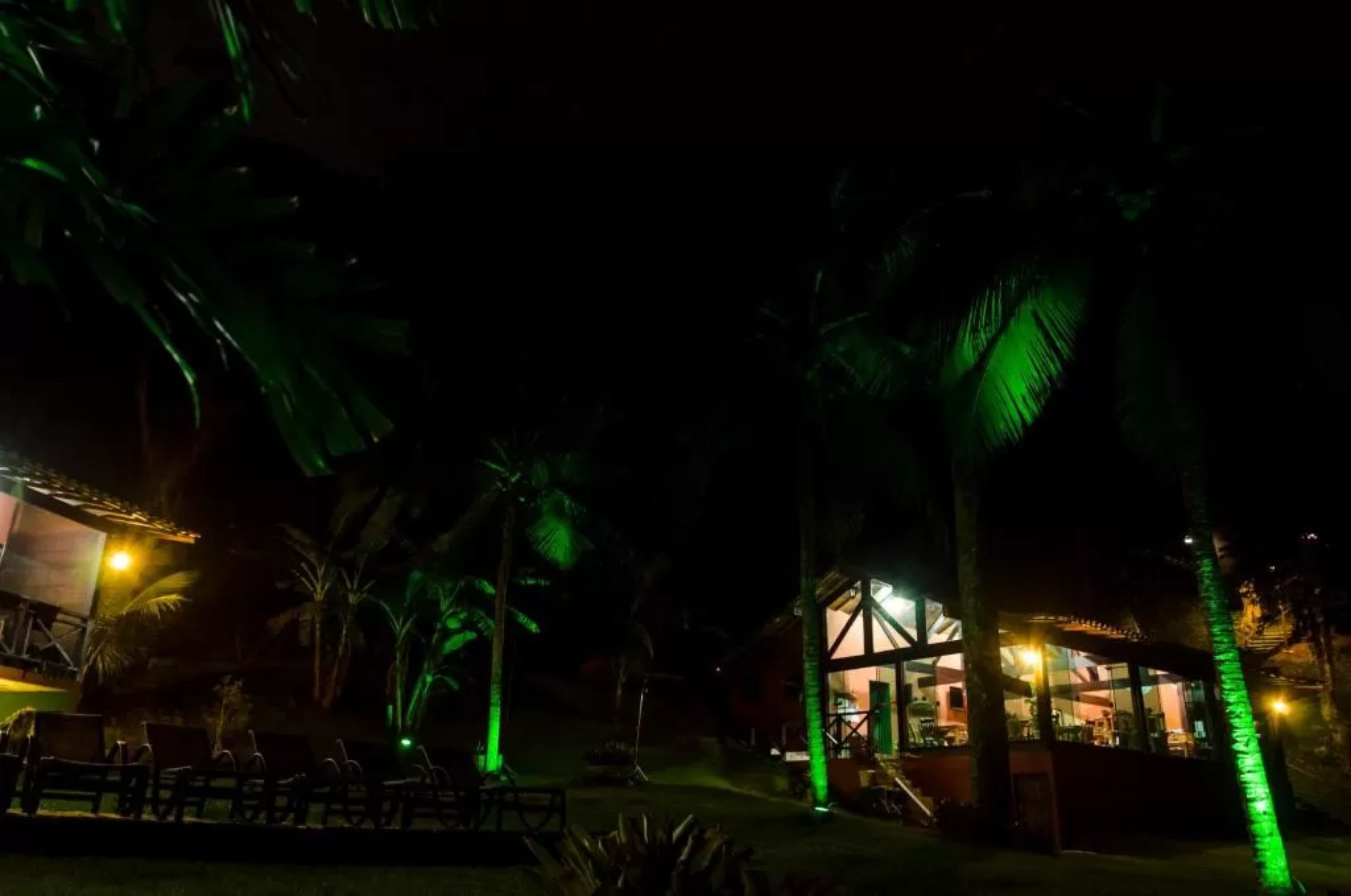Vista noturna de um resort com área de piscina, palmeiras e um edifício com luzes verdes iluminadas.
