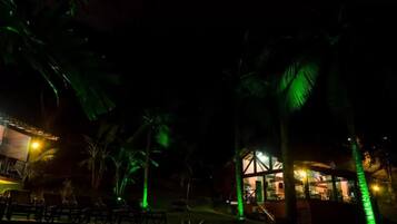 Vista noturna de um resort com área de piscina, palmeiras e um edifício com luzes verdes iluminadas.
