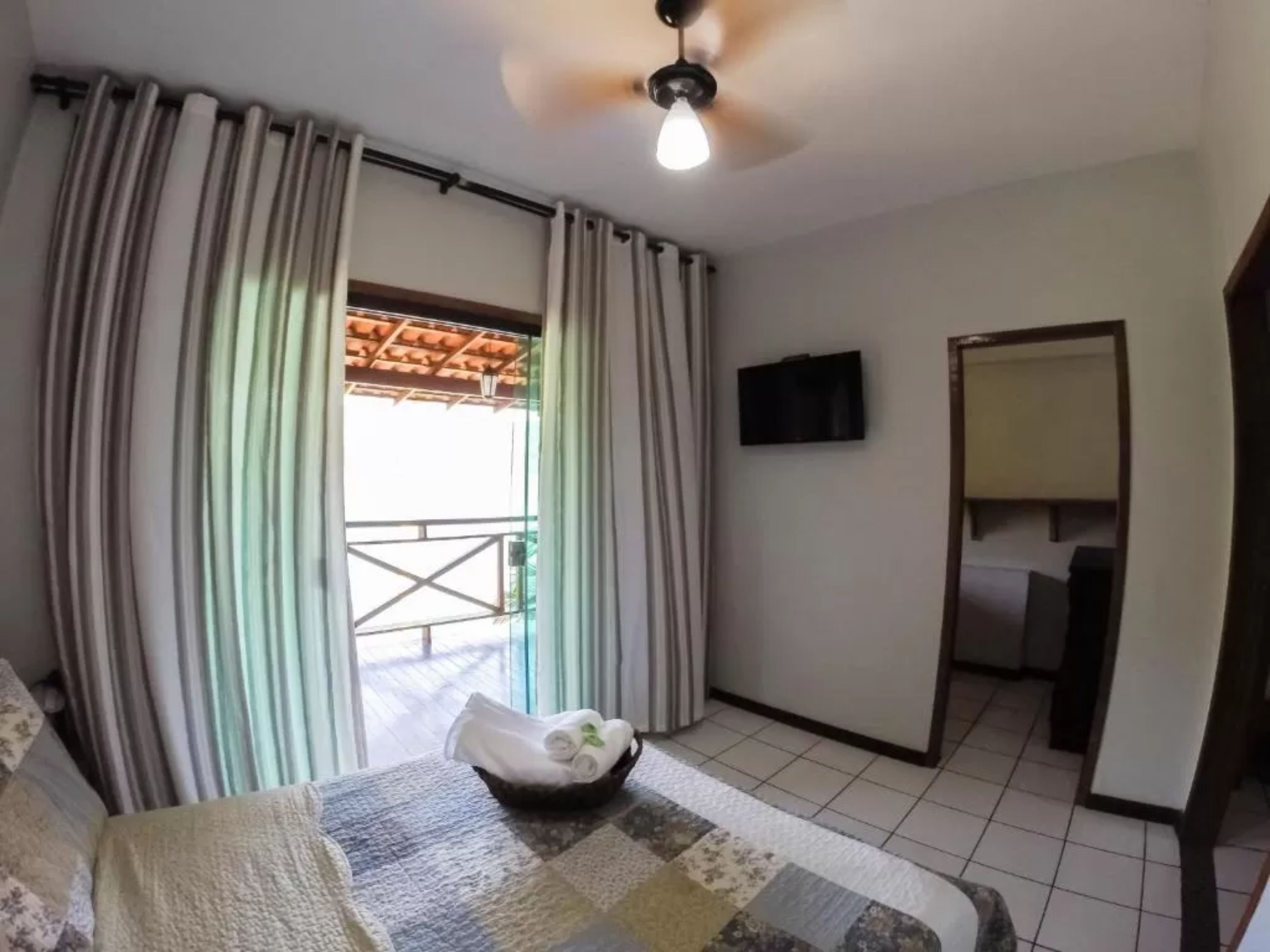 Quarto de hotel com cama, ventilador de teto, televisão e vista para a varanda externa.