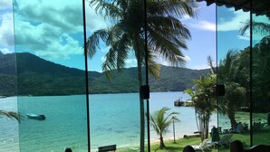 A view from a room with large windows overlooking a lake, with a boat on the water and a dock with people. - Pousada Portal do Sol (Angra dos Reis)