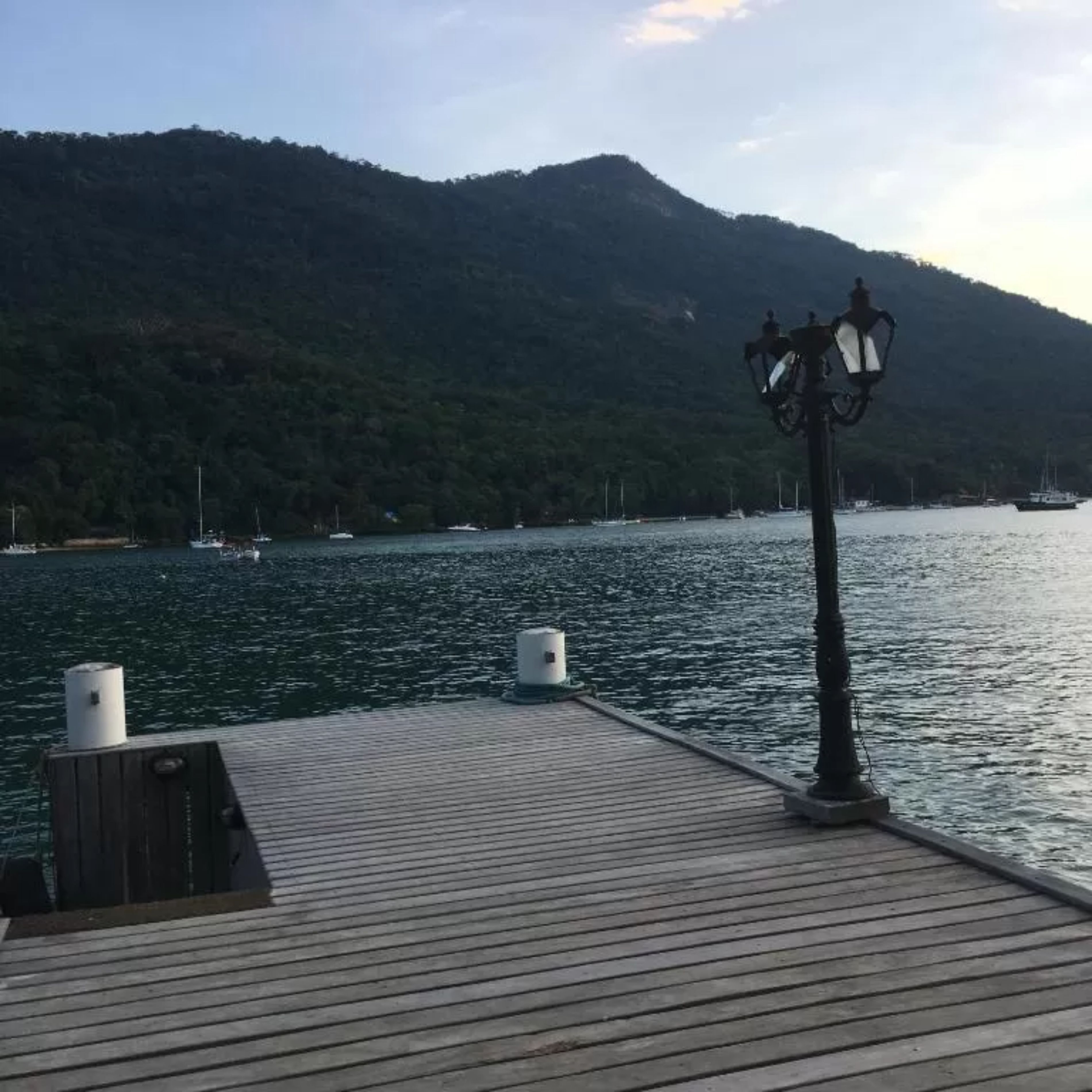 A wooden dock with a lamp post and two bollards, overlooking a calm body of water with boats and a forested hill in the background.