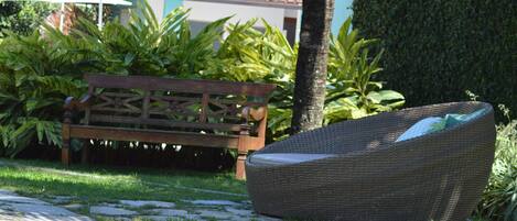 A wicker lounge chair and a wooden bench in a garden setting with a tiled-roof building in the background.