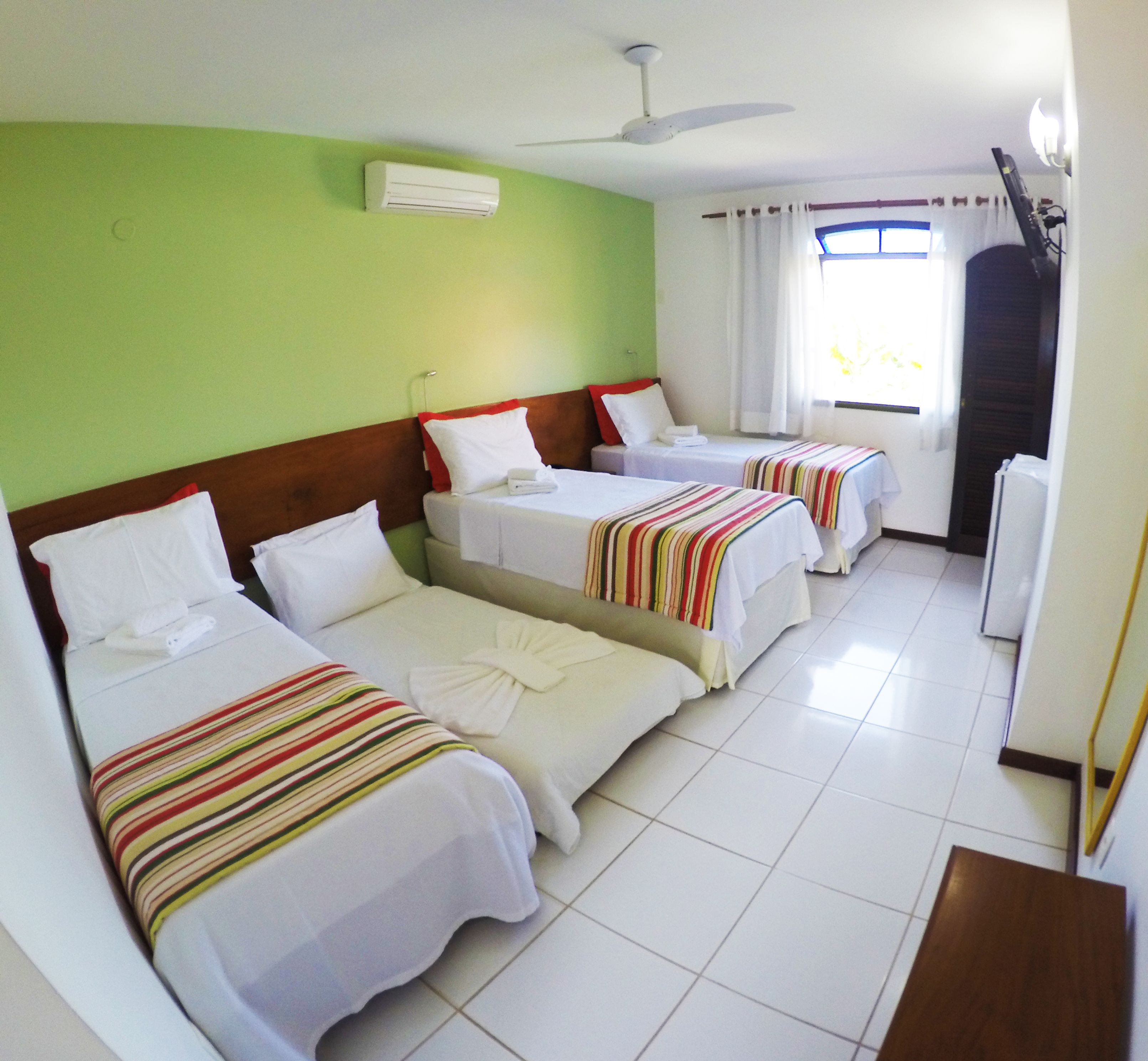 Habitación de hotel con tres camas individuales, ventilador de techo, aire acondicionado y ventana con cortinas.