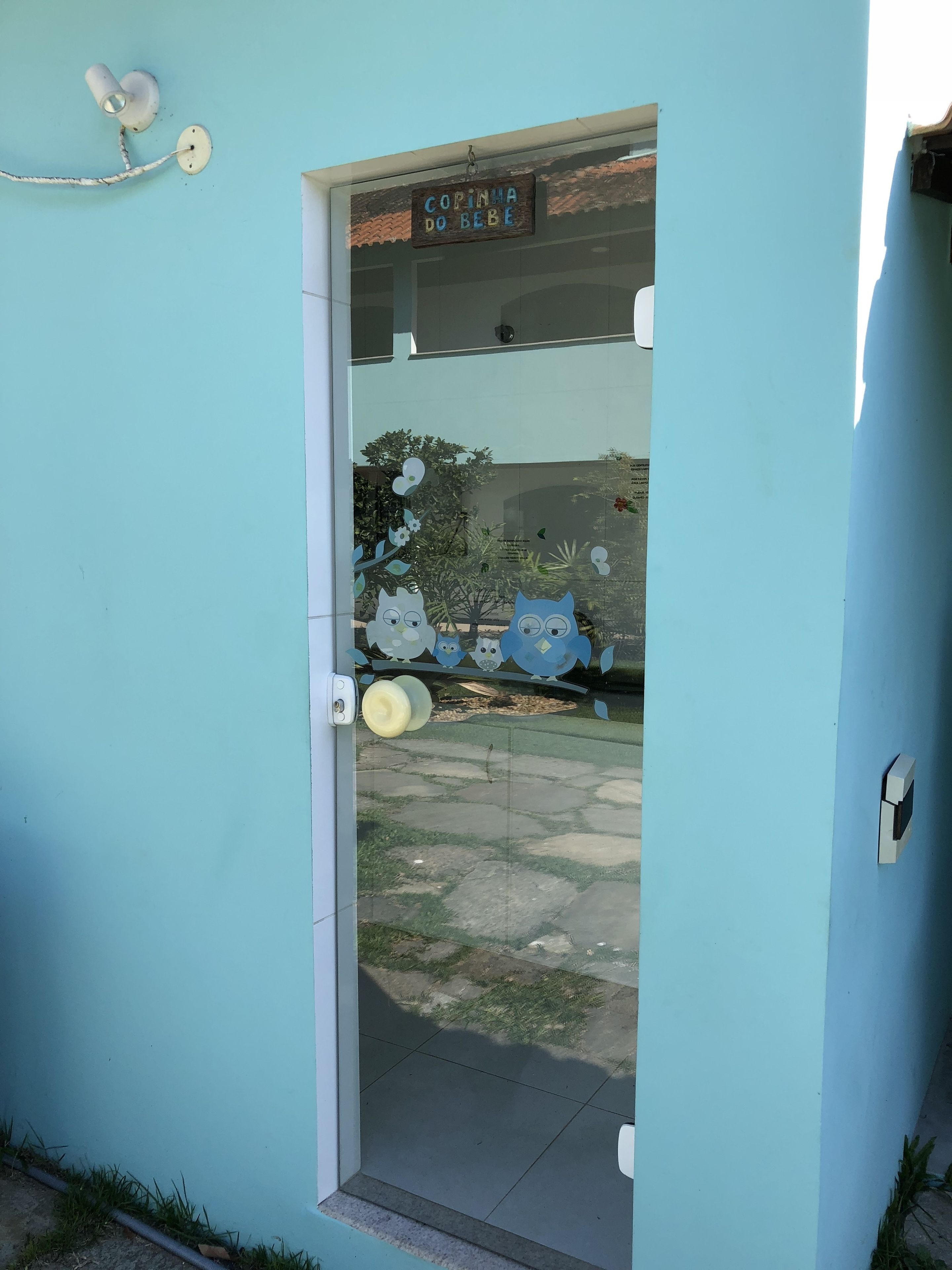 Una puerta con un letrero que dice 'Caminha do Bebê' (paseo para bebés), una puerta de cristal con adornos de lechuzas y un pequeño escalón que da acceso a un edificio.
