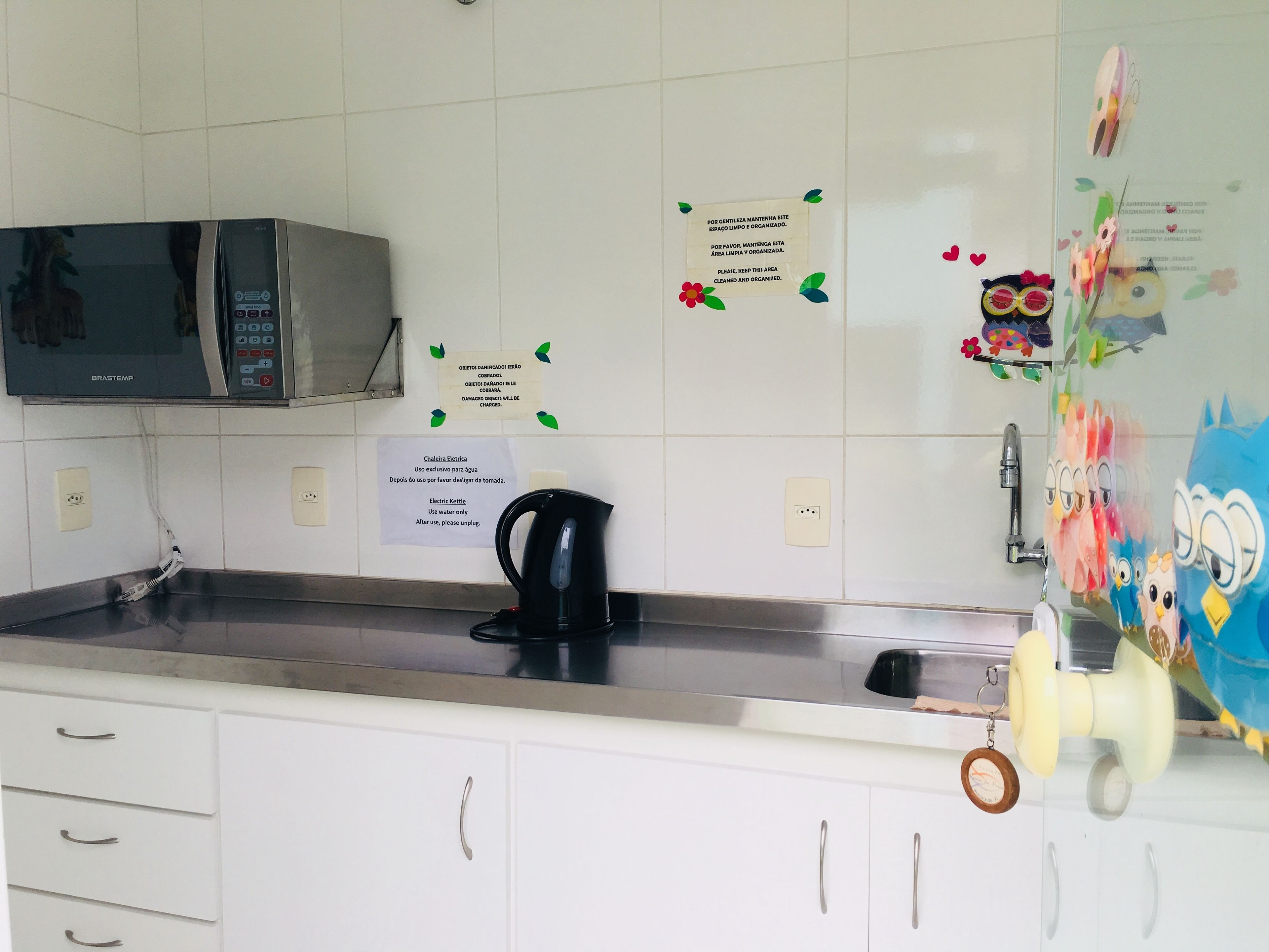 A kitchen with white cabinets, a microwave, a kettle, and a refrigerator with children's drawings.