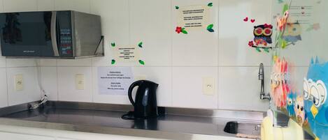 A kitchen with white cabinets, a microwave, a kettle, and a refrigerator with children's drawings.