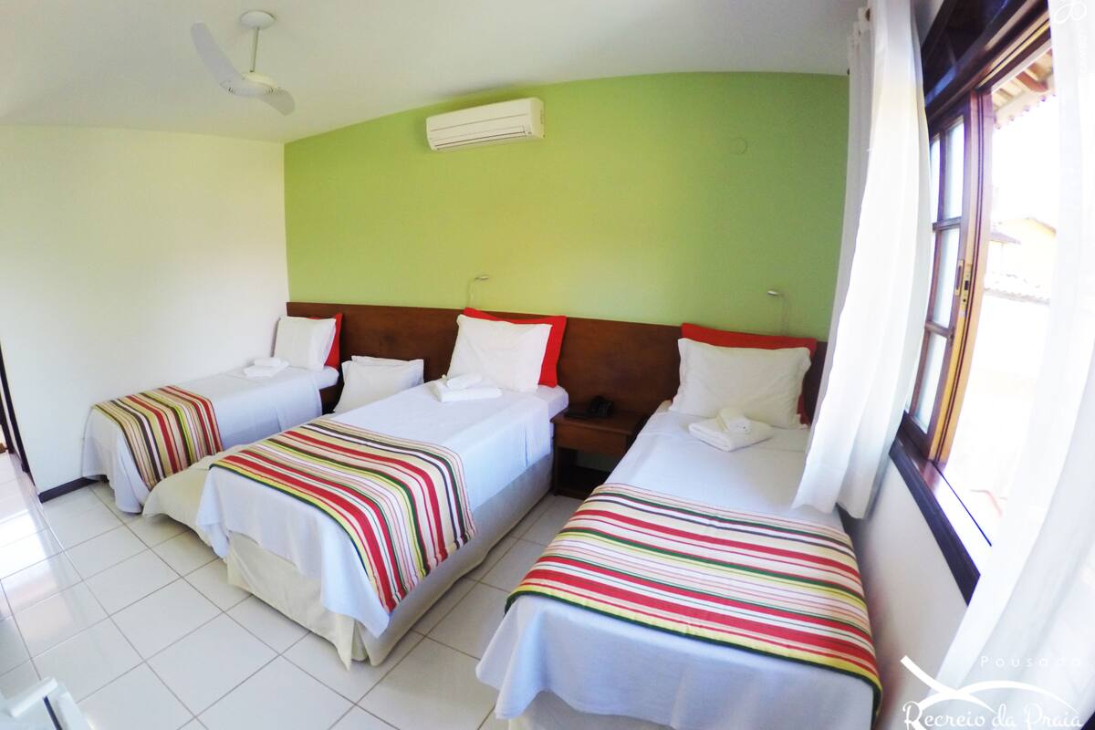 Quarto de hotel com três camas, cada uma com um cobertor listrado, uma mesa de cabeceira e uma janela com cortinas.