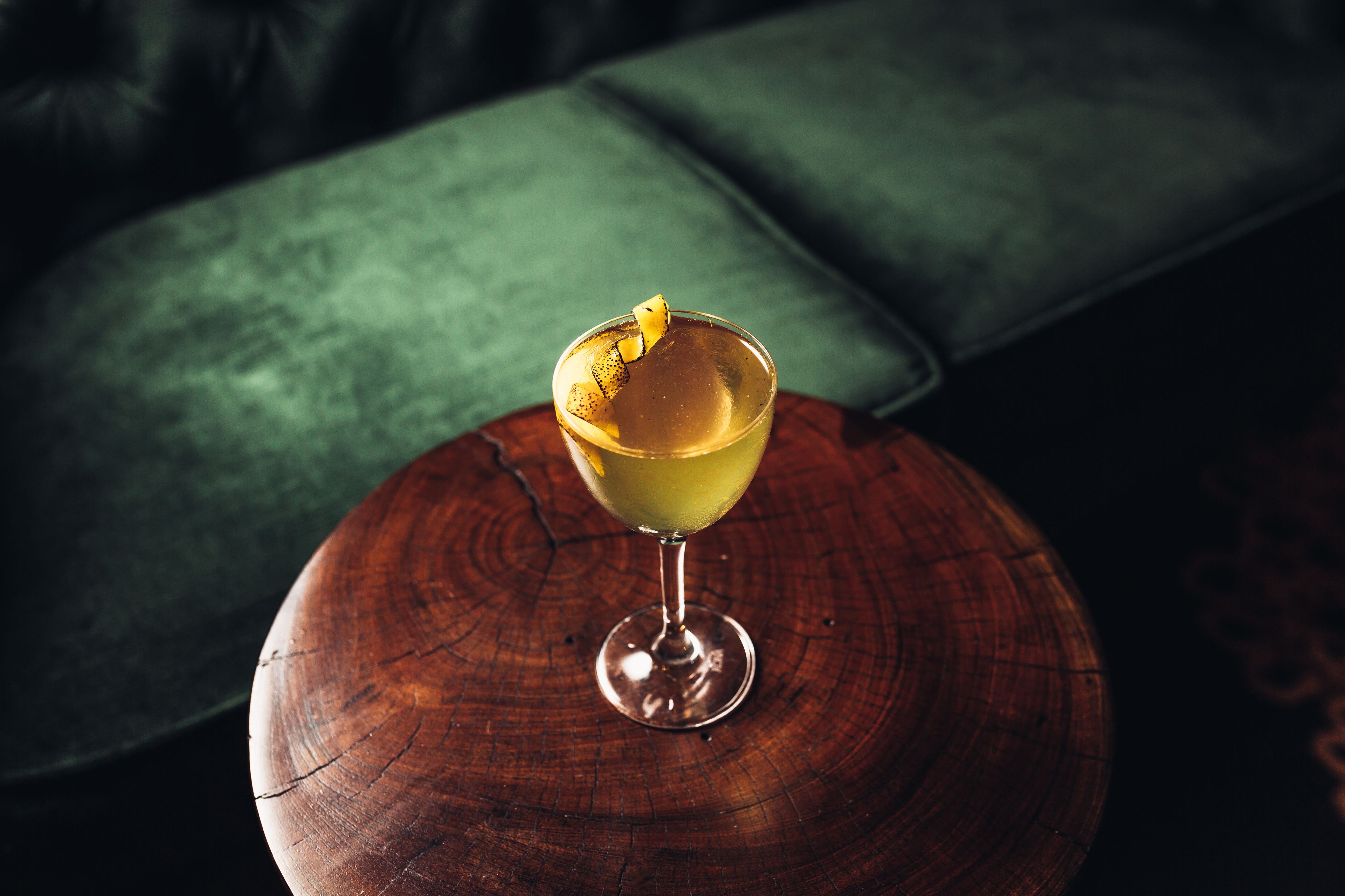 A cocktail on a wooden coaster with a green upholstered sofa in the background.