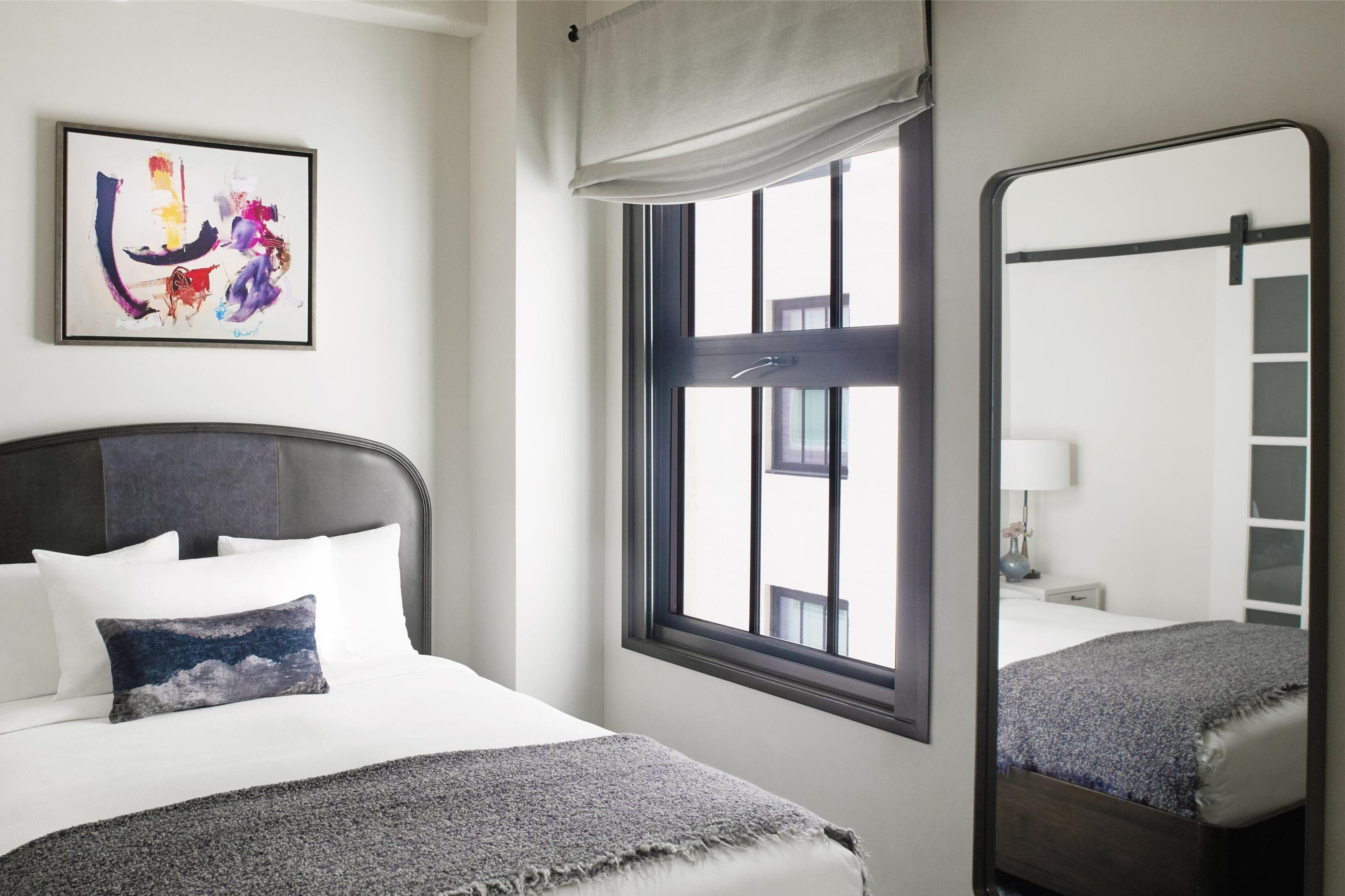 A hotel room with a bed, a window with a view, a mirror, and a painting on the wall.