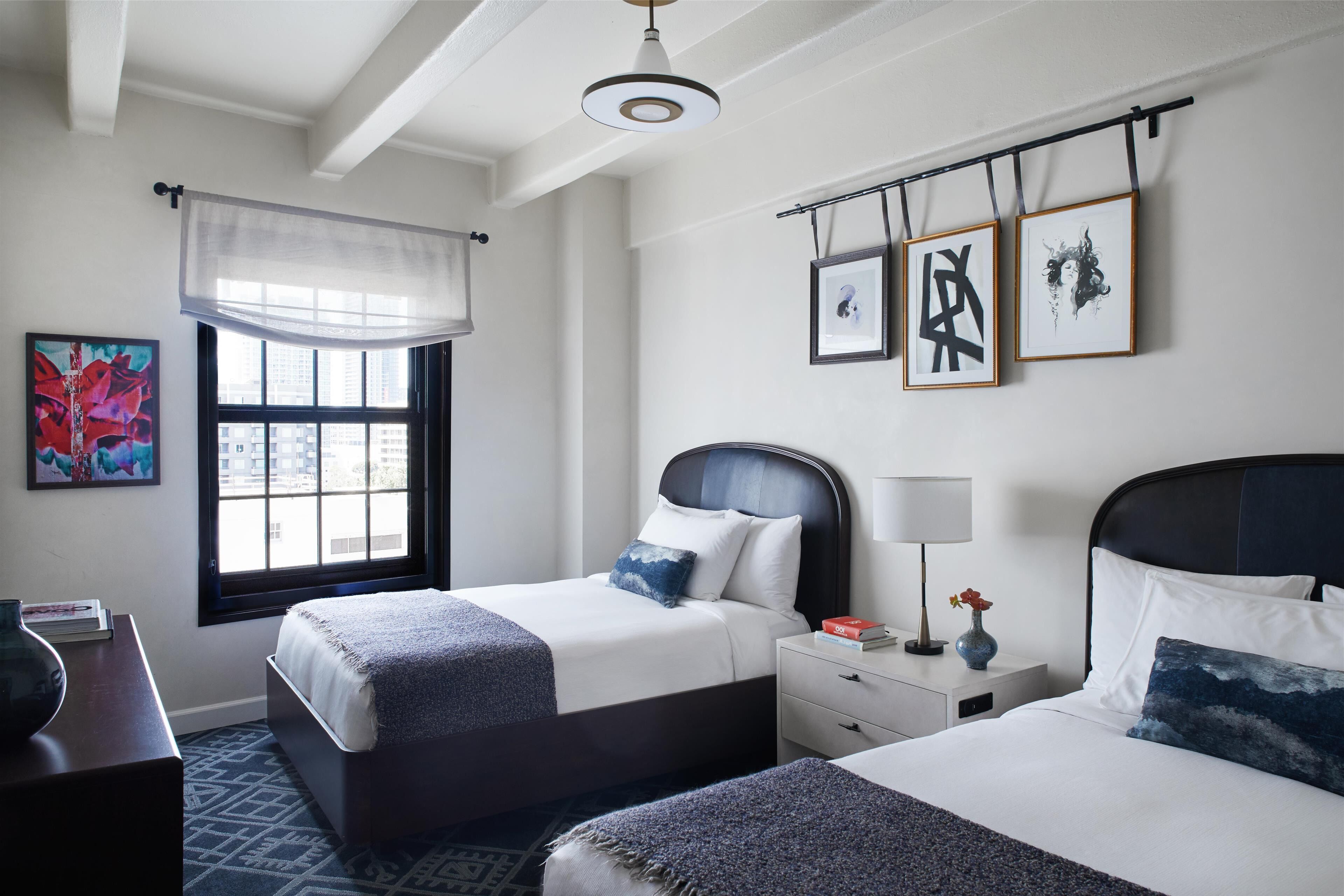 A hotel room with two beds, a large window with a view of buildings, a ceiling fan, and artwork on the walls.