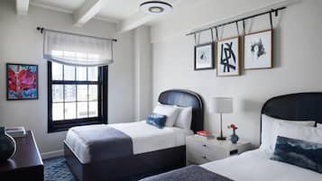 A hotel room with two beds, a large window with a view of buildings, a ceiling fan, and artwork on the walls.