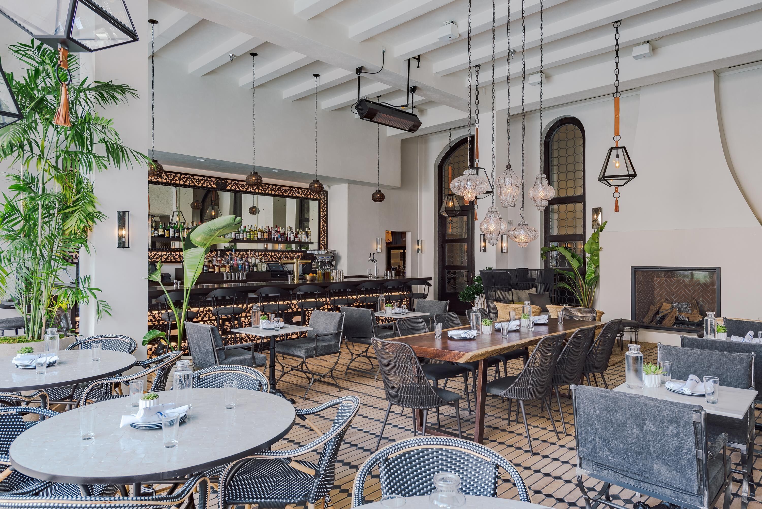 A restaurant with round tables, black and white wicker chairs, hanging pendant lights, and a bar area with shelves of bottles.