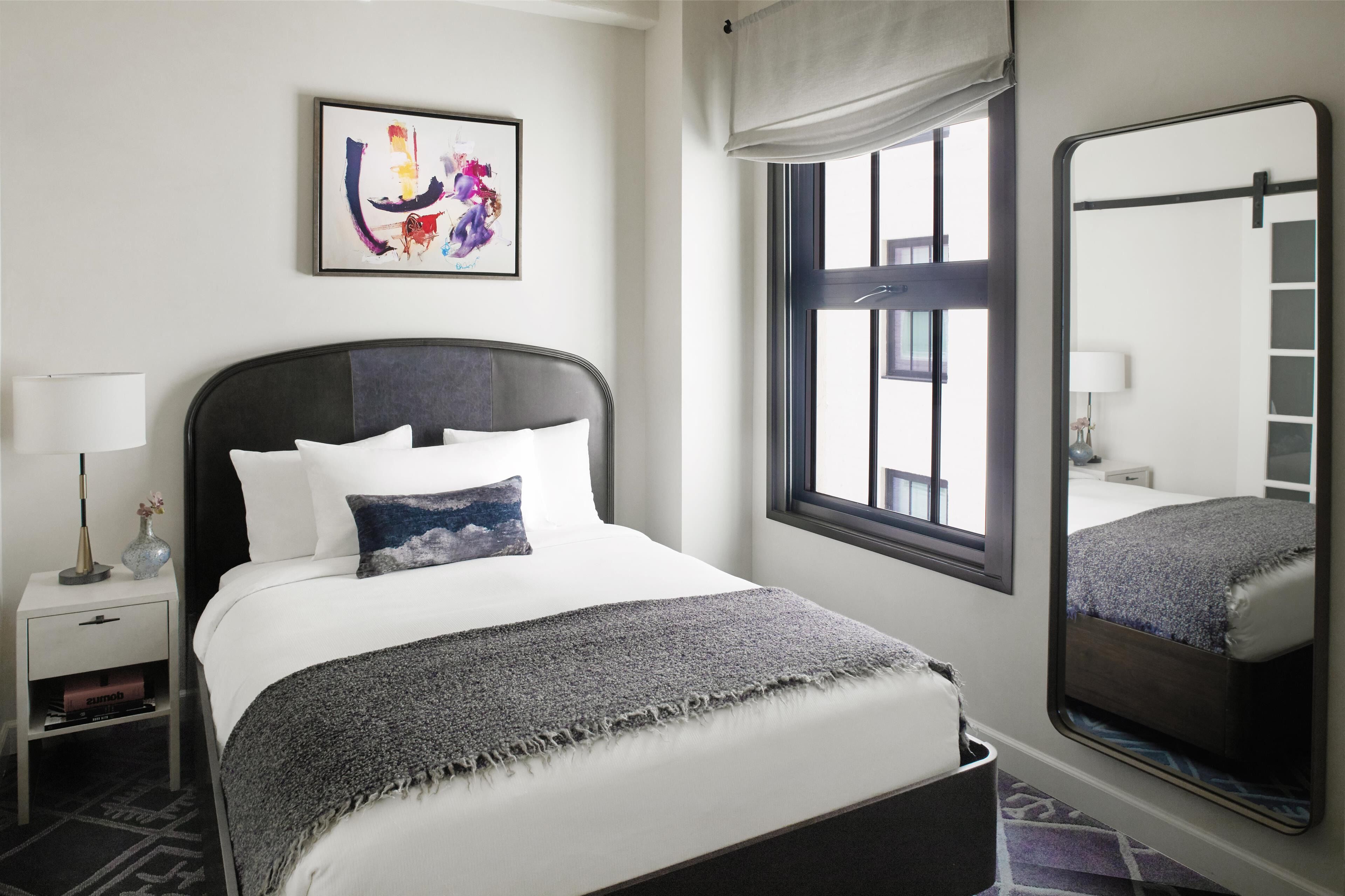 A neatly arranged hotel room with a bed, a bedside table, a lamp, a framed artwork, a window with a view, and a mirror.