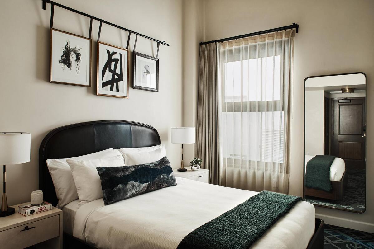 Ein ordentlich eingerichtetes Hotelzimmer mit einem großen Bett, Nachttischlampen, einem Nachttisch, einem gerahmten Kunstwerk über dem Bett, einem Fenster mit Vorhängen und einem Spiegel an der Wand.