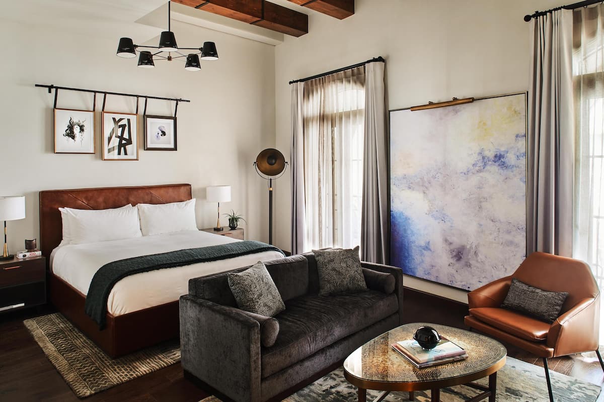 Ein modernes Hotelzimmer mit einem großen Bett, einer Couch, einem Couchtisch und einem Sessel. An den Wänden befinden sich Bilder in Rahmen, eine Stehlampe und ein großes abstraktes Gemälde.