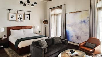 Ein modernes Hotelzimmer mit einem großen Bett, einer Couch, einem Couchtisch und einem Sessel. An den Wänden befinden sich Bilder in Rahmen, eine Stehlampe und ein großes abstraktes Gemälde.