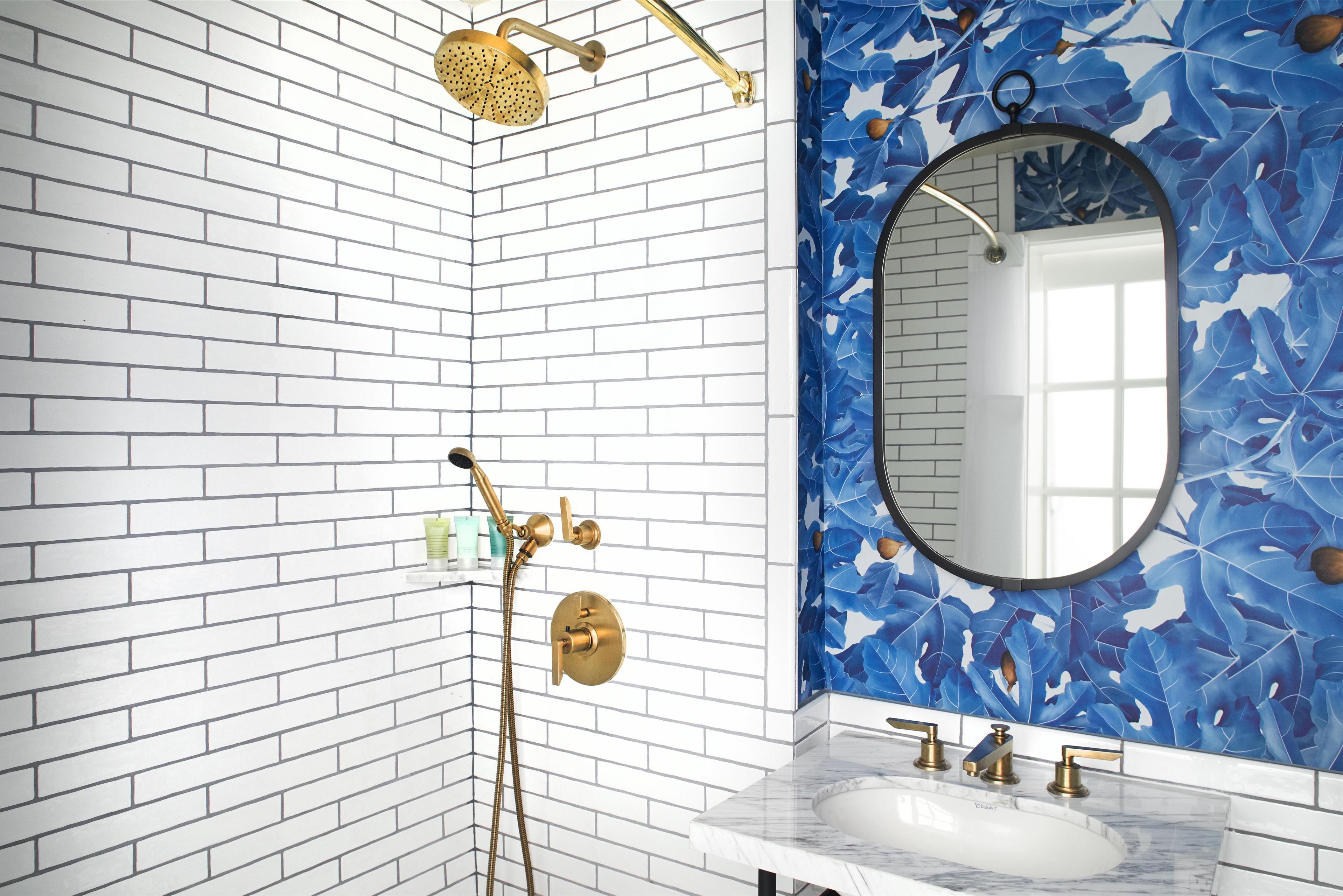 Une salle de bain avec des murs en briques blanches, un mur orné d’un motif de feuilles bleues, un miroir rond, un lavabo blanc et des accessoires dorés.