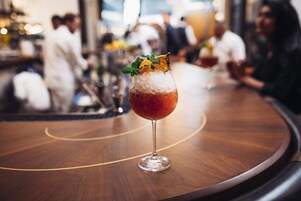 Een bar met een ronde houten tafel en een glas rode drank, versierd met munt en een sinaasappelschijfje.