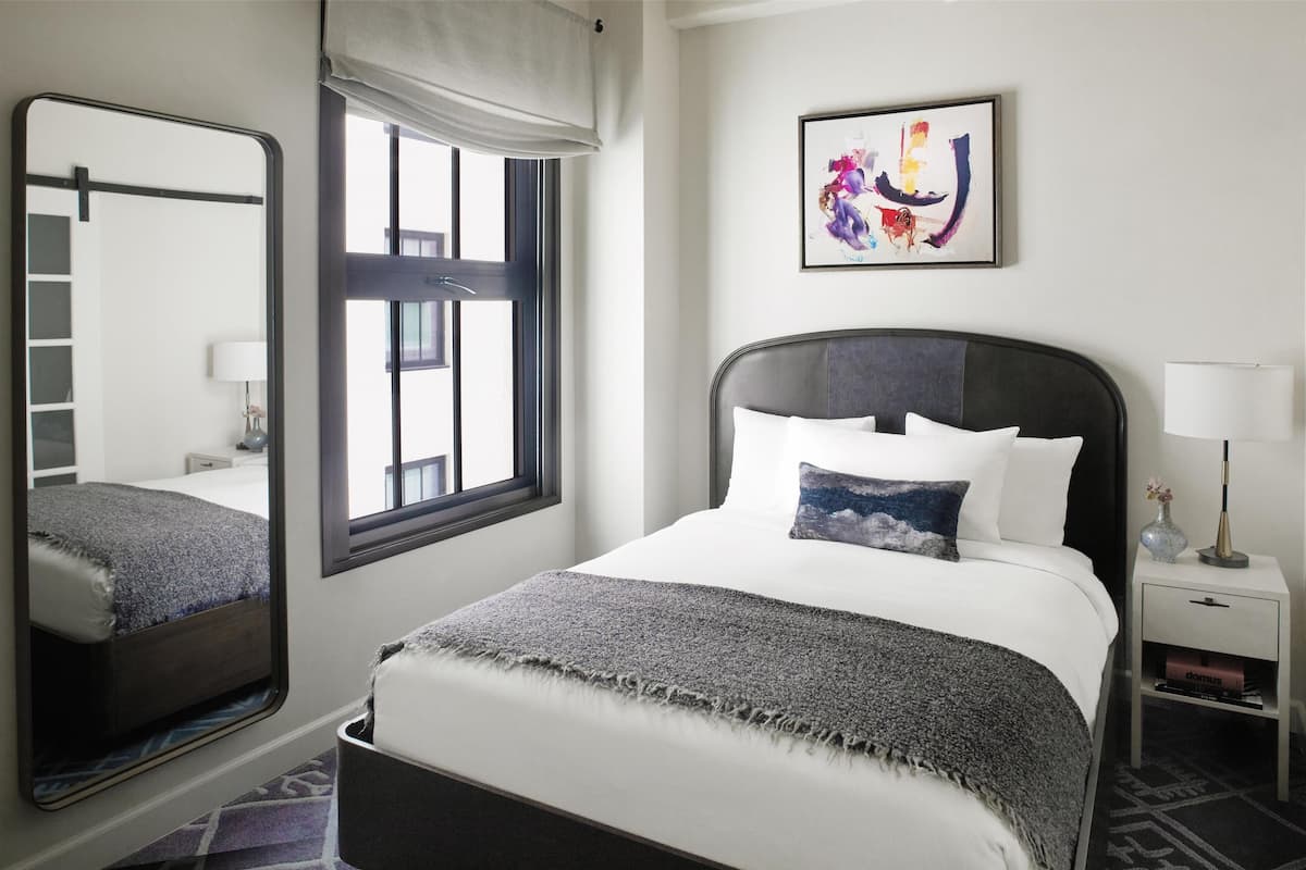 Ein ordentlich eingerichtetes Hotelzimmer mit einem großen Bett, einem Nachttisch mit Lampe, einem Fenster mit Blick und einem Spiegel, der den Raum wiedergibt.