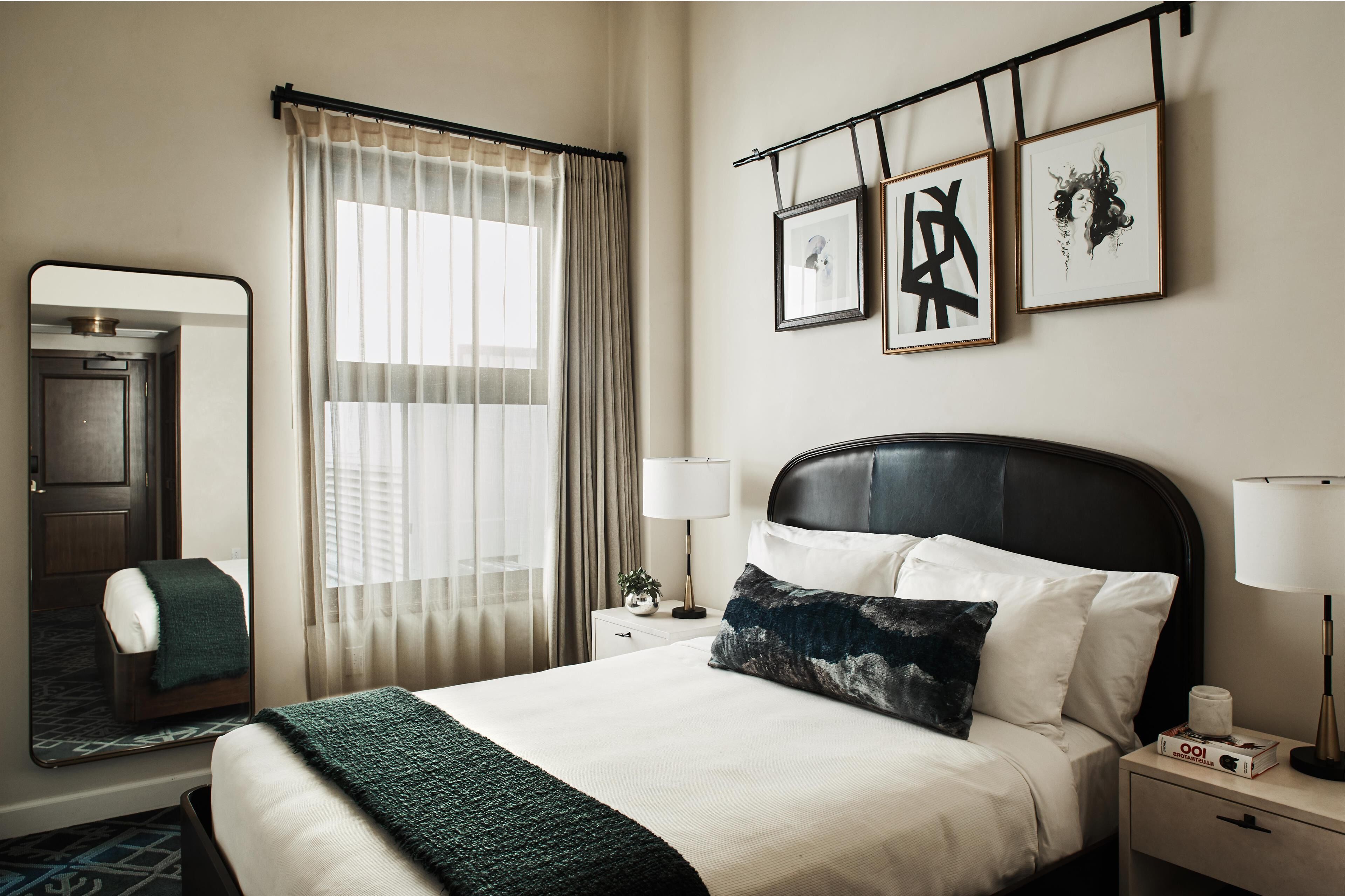 Ein ordentlich eingerichtetes Hotelzimmer mit einem großen Bett, zwei Nachttischen, einem Spiegel, einem Fenster mit Vorhängen und Kunstwerken an der Wand.