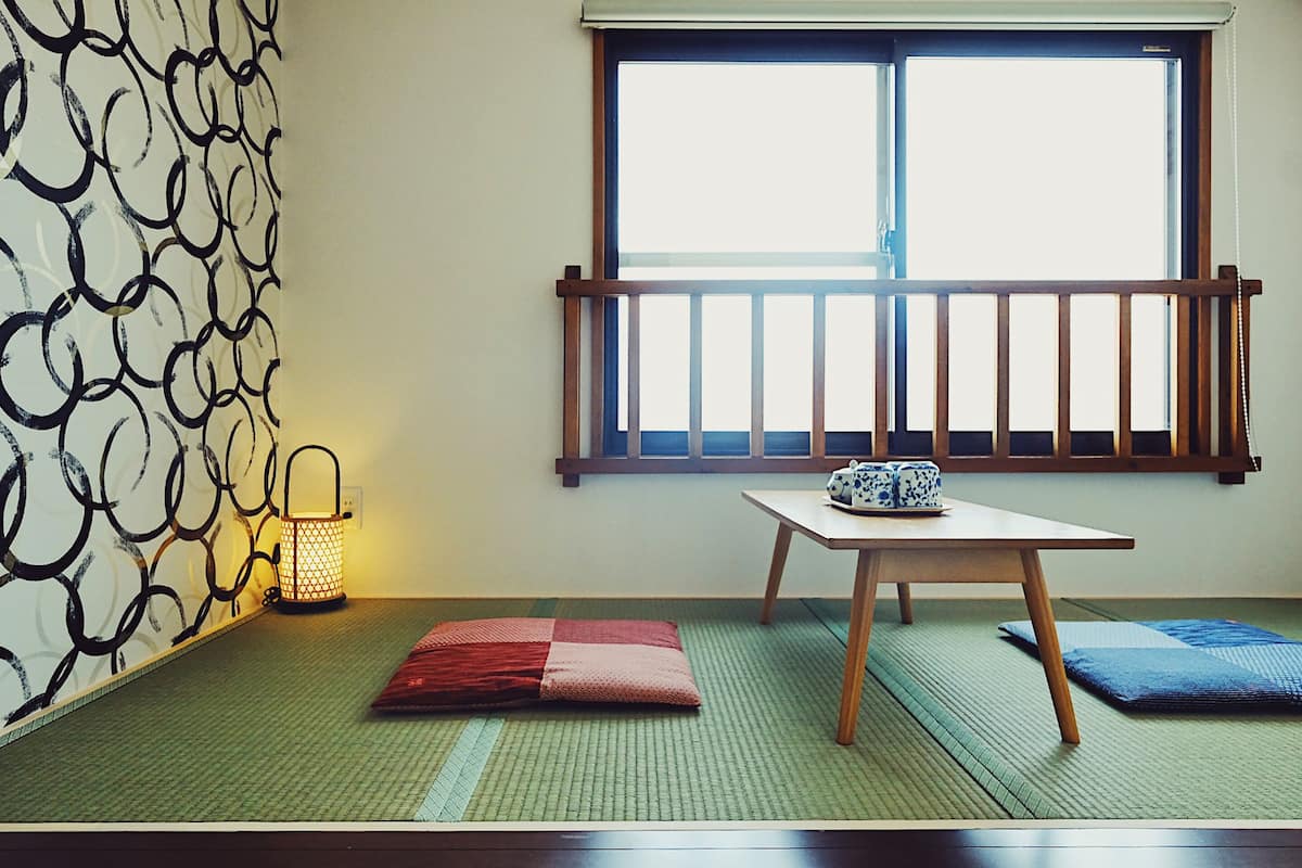 A room with tatami mats, a low table with a ceramic pot, a hanging lantern, and a window with a wooden blind.