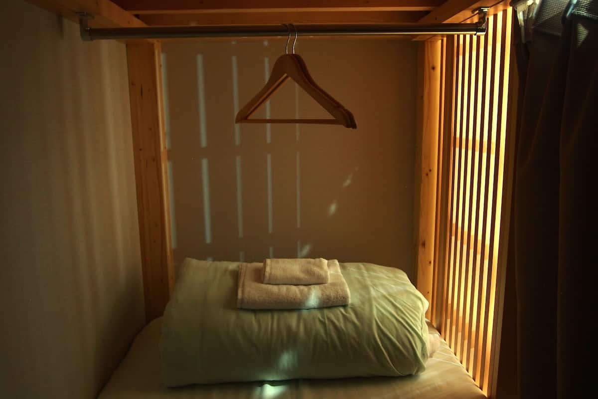 A small, cozy room with a bed, a wooden hanger, and a window with vertical blinds.