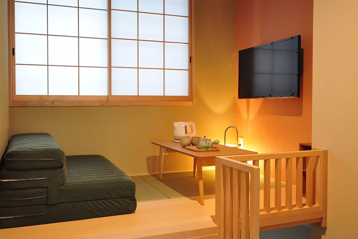 A compact room with a futon bed, a small table with a kettle and cups, a television mounted on the wall, and a window with a grid pattern.