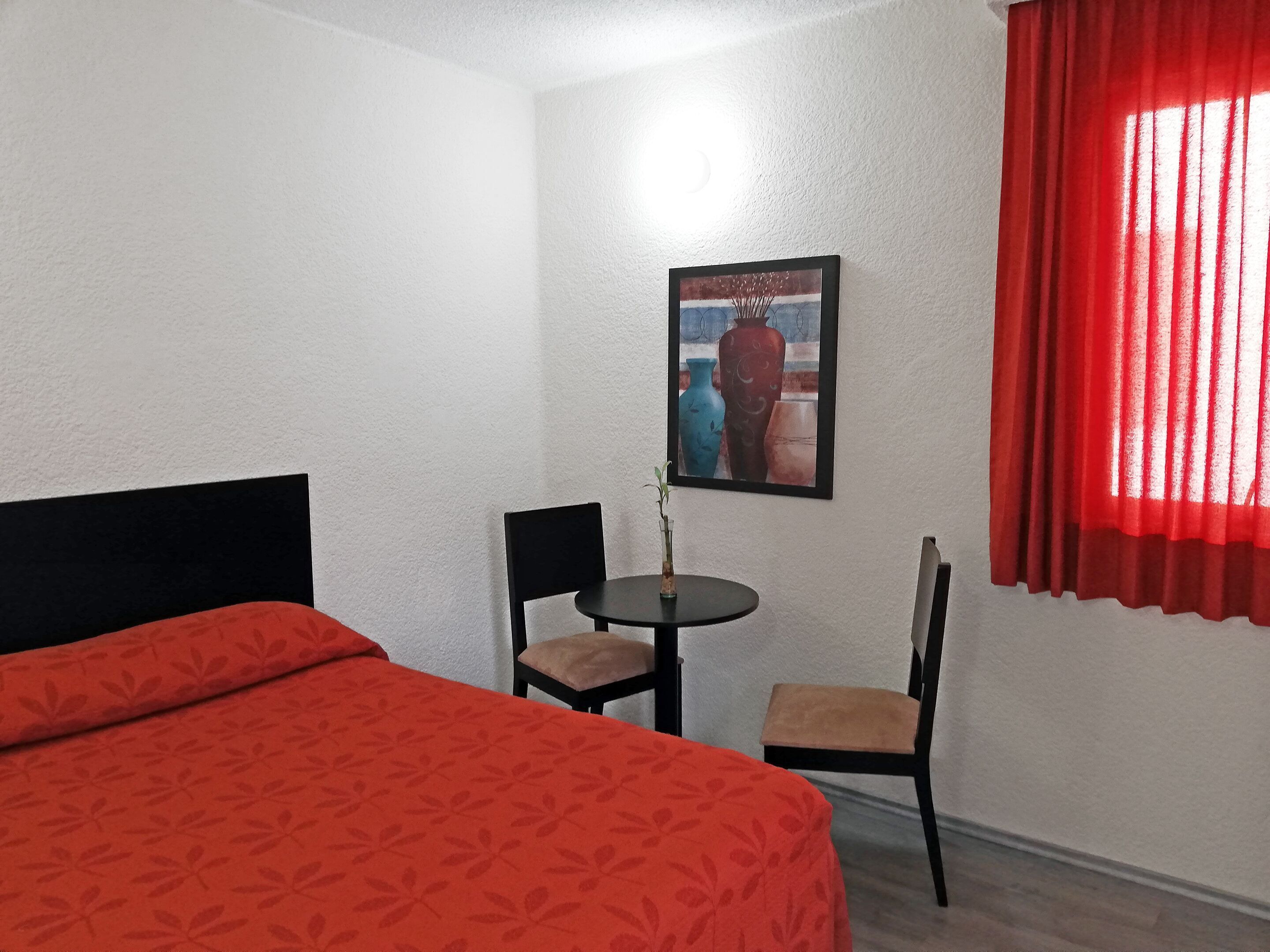 A hotel room with a bed, two chairs, a small table with a vase, and a painting on the wall.