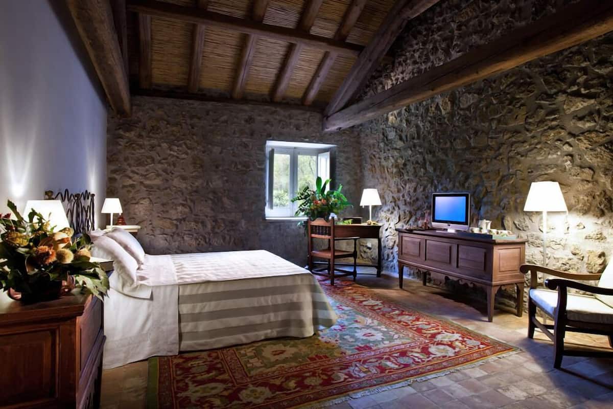 Una habitación rústica con pared de piedra, una cama con colcha estampada, un escritorio con televisor y una ventana con luz natural.
