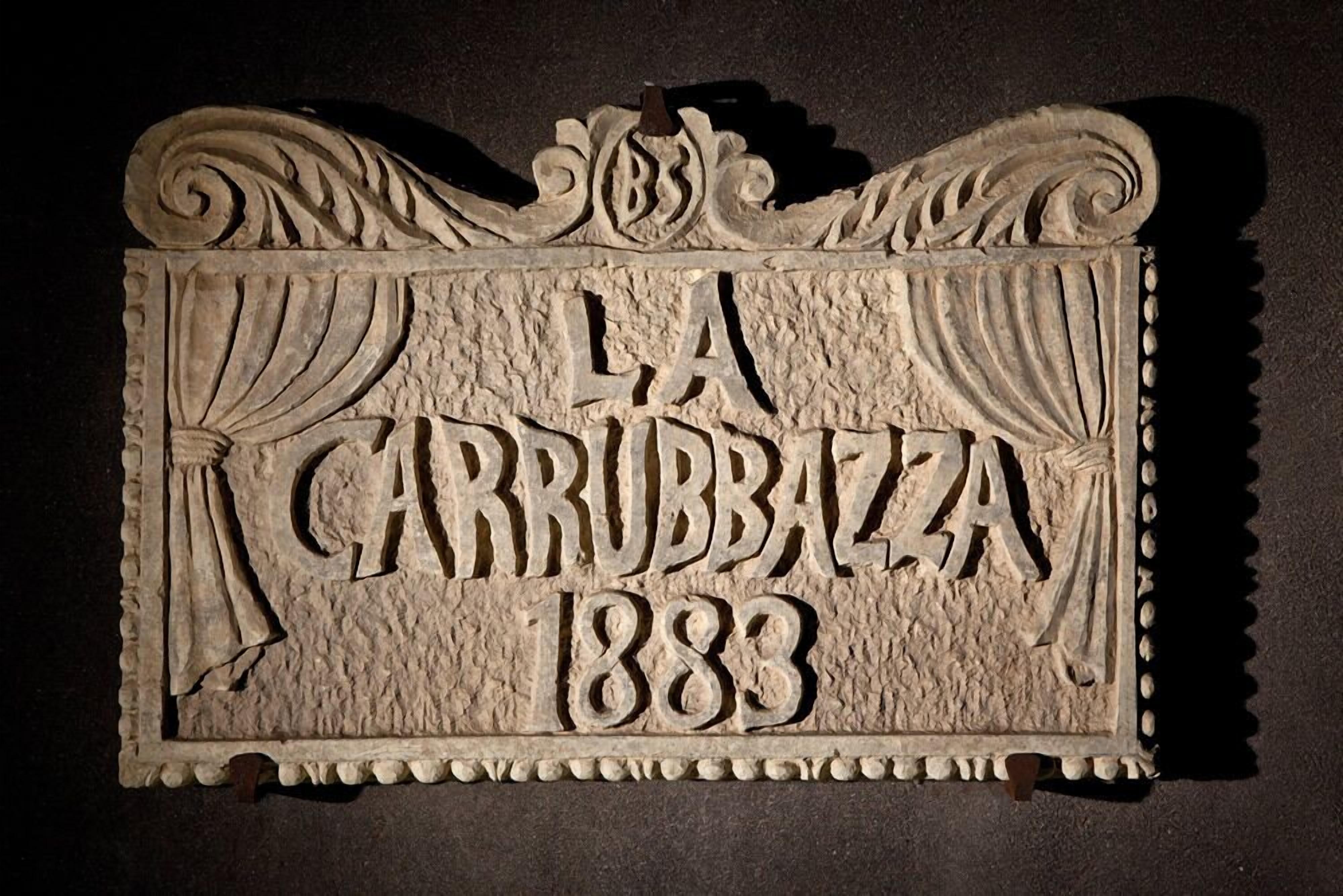 Un senyal tallat amb el text "LA CARROZZA 1883" i elements decoratius com cortines i ornaments en forma de volutes.