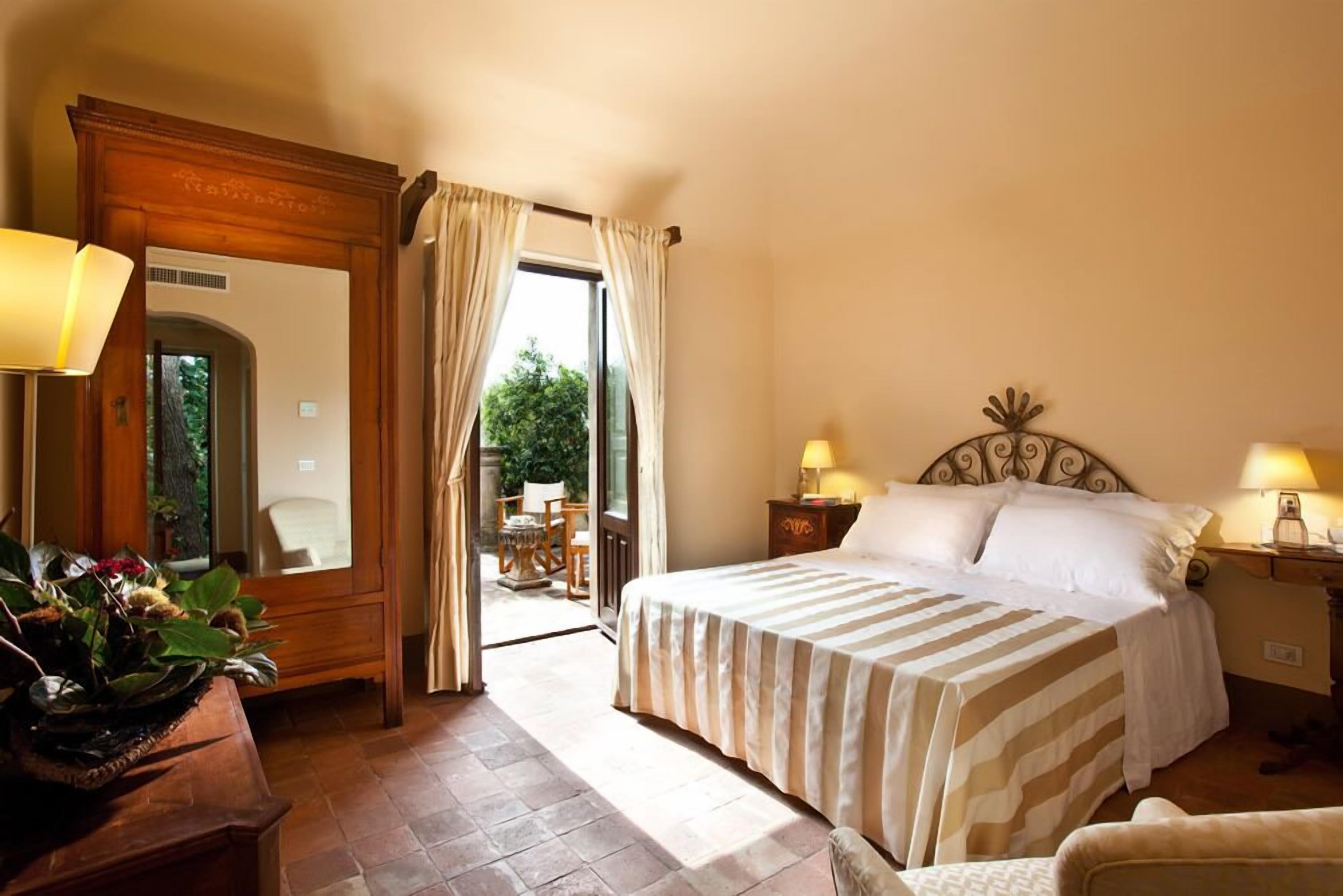 Une chambre d’hôtel avec un grand lit, deux lampes de chevet, une tête de lit en bois et une vue sur un espace extérieur, accessible par une porte ouverte.