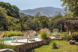 Zona de piscina con sillas de descanso, una estructura de techo de paja y montañas al fondo.