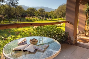 Una mesa redonda de vidrio con libros y un cuenco de fruta, situada en un balcón con vistas a un jardín exuberante y montañas lejanas.