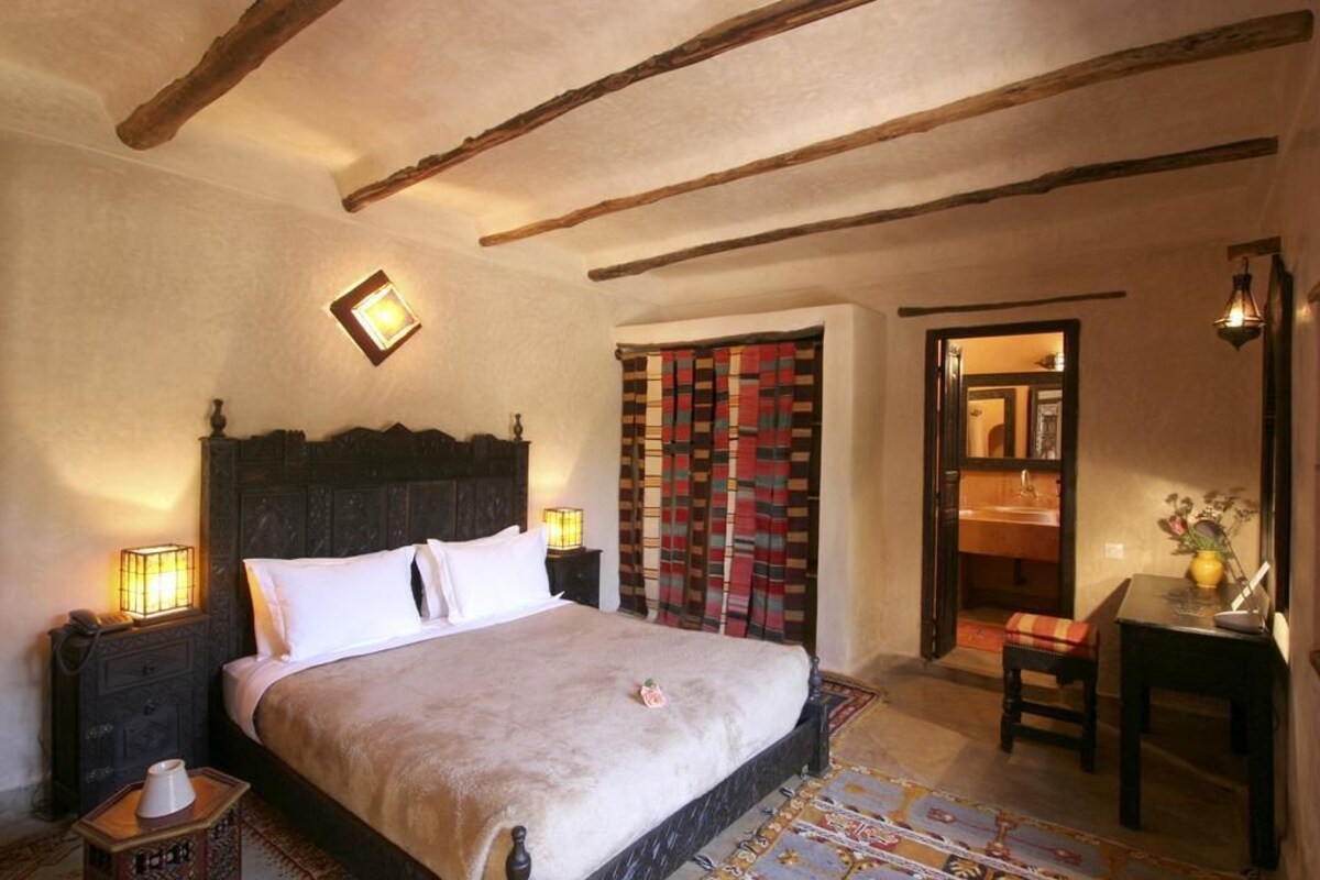 A bedroom with a large bed, wooden beams on the ceiling, a nightstand with a lamp, and a view into the bathroom.