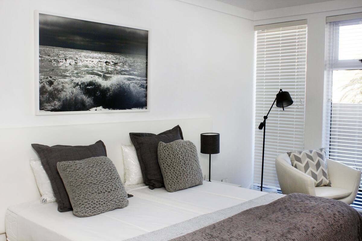 Ein Schlafzimmer mit einem Bett, Kissen, einer Lampe, einem Sessel und einem großen gerahmten Bild an der Wand.