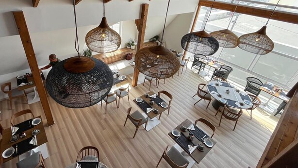 Een ruime eetruimte met hoge plafonds en grote ramen, met ronde tafels en hanglampen van riet.