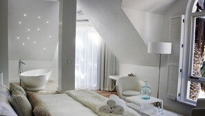 Een moderne slaapkamer met een groot bed, een witte stoel, een klein tafeltje en een groot raam met uitzicht op bomen.