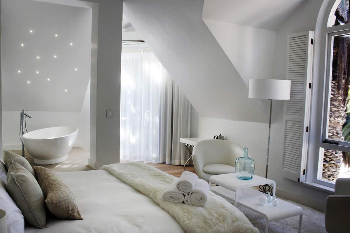 Ein modernes Schlafzimmer mit einem großen Bett, einem weißen Stuhl, einem kleinen Tisch und einem großen Fenster mit Blick auf Bäume.