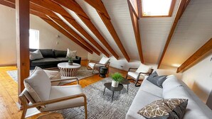 Een ruime loft met hoge plafonds, zichtbare houten balken, een gezellige zithoek met een bank, stoelen en een salontafel, en een groot dakraam dat natuurlijk licht binnenlaat.