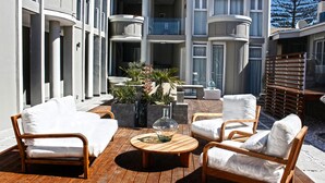 Een zonnig terras met een houten tafel, witte stoelen met kussens en potplanten.