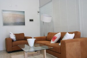 Ein modernes Wohnzimmer mit zwei braunen Sofas, einem Glastisch, einer weißen Vase und einem großen abstrakten Gemälde an der Wand.