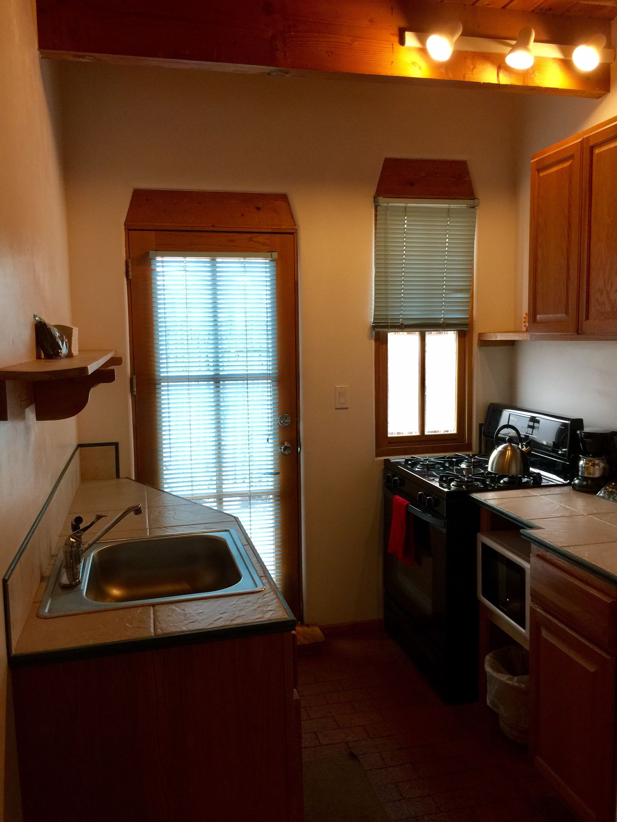 A compact kitchen with a sink, stove, and oven. There is a window with blinds, a wooden door, and a shelf above the sink.