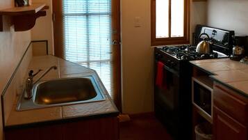 A compact kitchen with a sink, stove, and oven. There is a window with blinds, a wooden door, and a shelf above the sink.